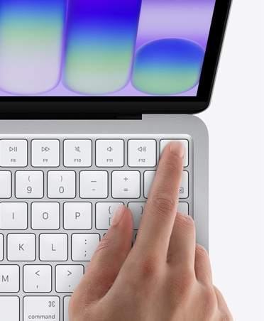 MacBook Neo sudraba krāsā, personas pirksts uz Touch ID sensora tastatūras augšējā labajā stūrī