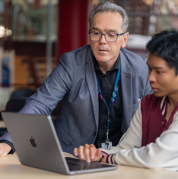 A man points to a laptop screen, while a student sitting at a table in a classroom looks on.
