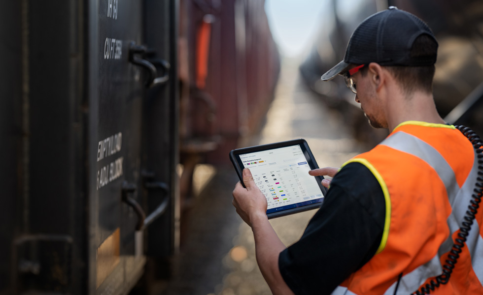 Un trabajador de pie junto a un tren revisa un iPad que muestra información de tráfico ferroviario