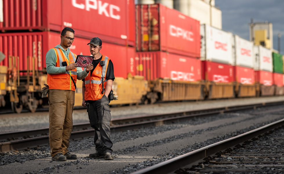 Dos trabajadores consultan un iPad de pie en un depósito ferroviario junto a un tren parado que transporta contenedores