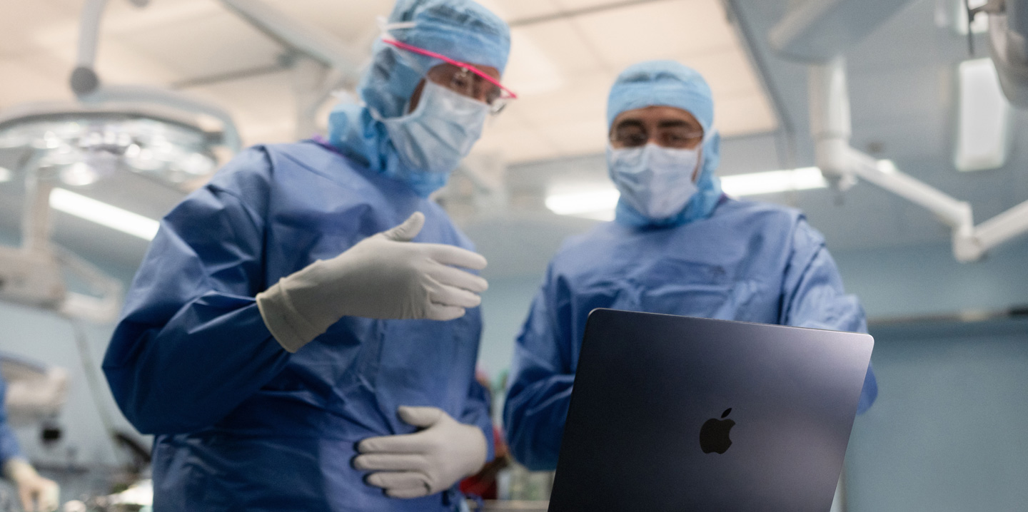 Dos cirujanos en el quirófano miran un MacBook, y uno hace gestos con la mano mientras el otro observa