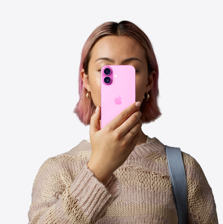 Persona pie sejas tur iPhone 16 ar rozā apdari, aizmugures skats augšējā stūrī pa kreisi atklāj divu kameru sistēmu.